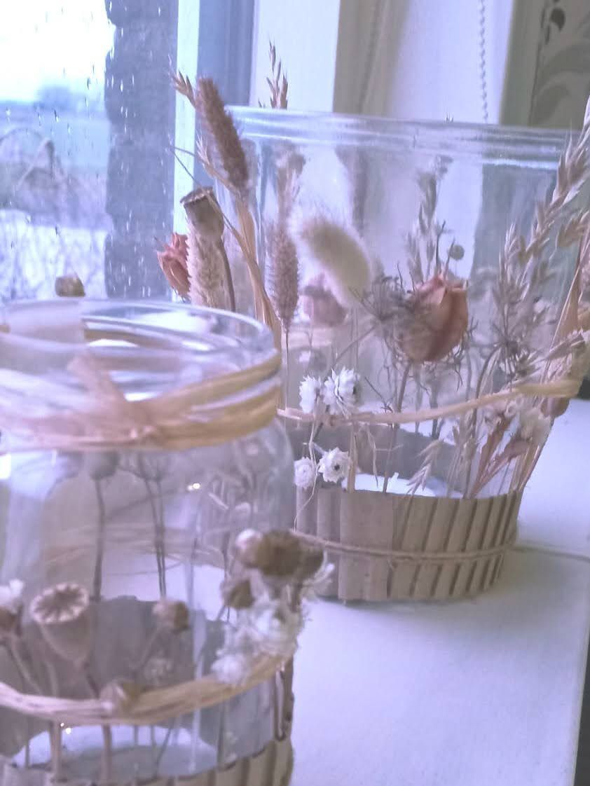 Twee glazen vazen met gedroogde bloemen bij een raam. De vazen zijn omwikkeld met touw en hout.