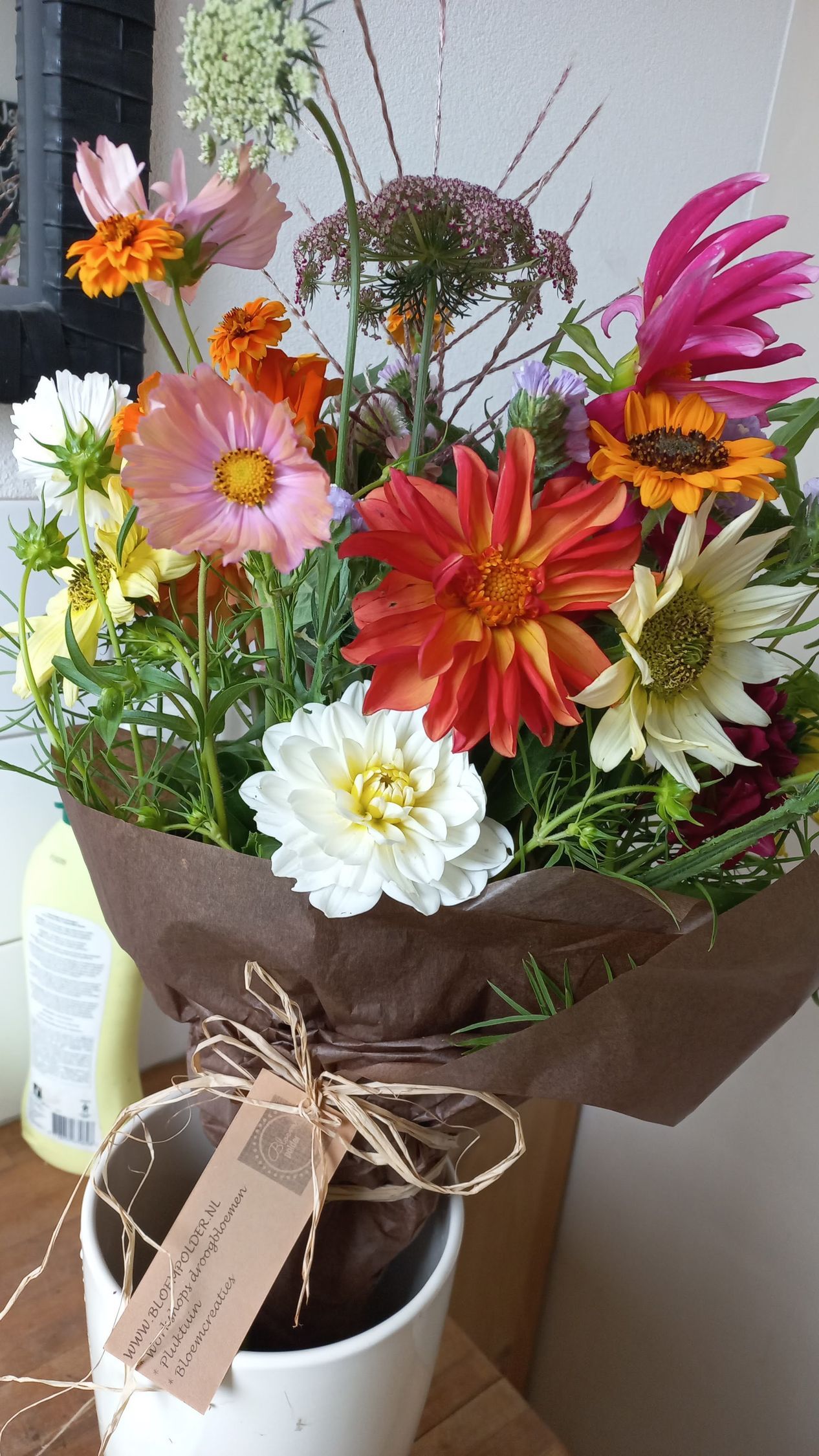Een boeket kleurrijke bloemen, verpakt in bruin papier en vastgebonden met touw, staat in een witte pot.
