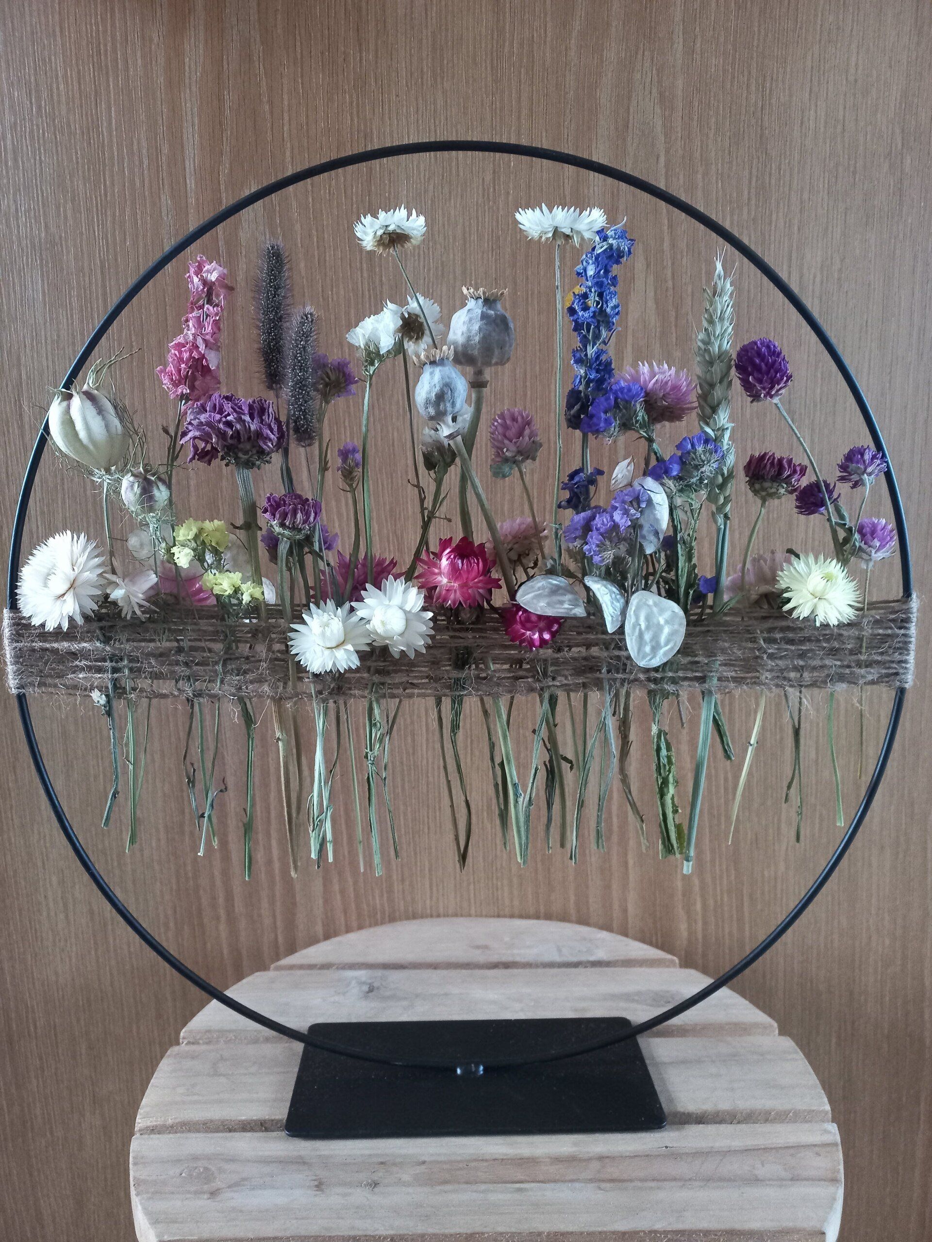 Een rond bloemstuk met diverse gedroogde bloemen, geplaatst op een standaard met een zwart tasje.