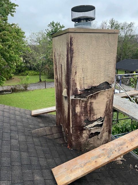Damaged concrete chimney with scorch marks and a bent cap beside a roof and yard.