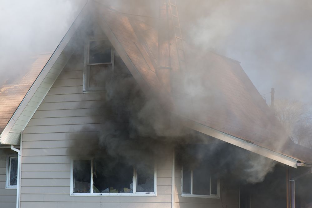Thick, dark smoke billows from the window and roof of a two-story residential house during a fire.