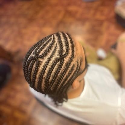 Close-up of braided cornrows on a person's head, wearing a white shirt, with a blurred indoor background