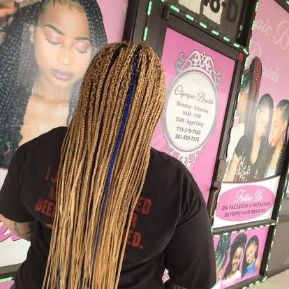 Person with long blonde braids standing before hair salon posters and signage.