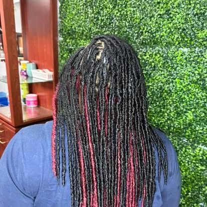 Back view of a person with black-and-red braids, seated indoors beside a wooden shelf and green wall paneling
