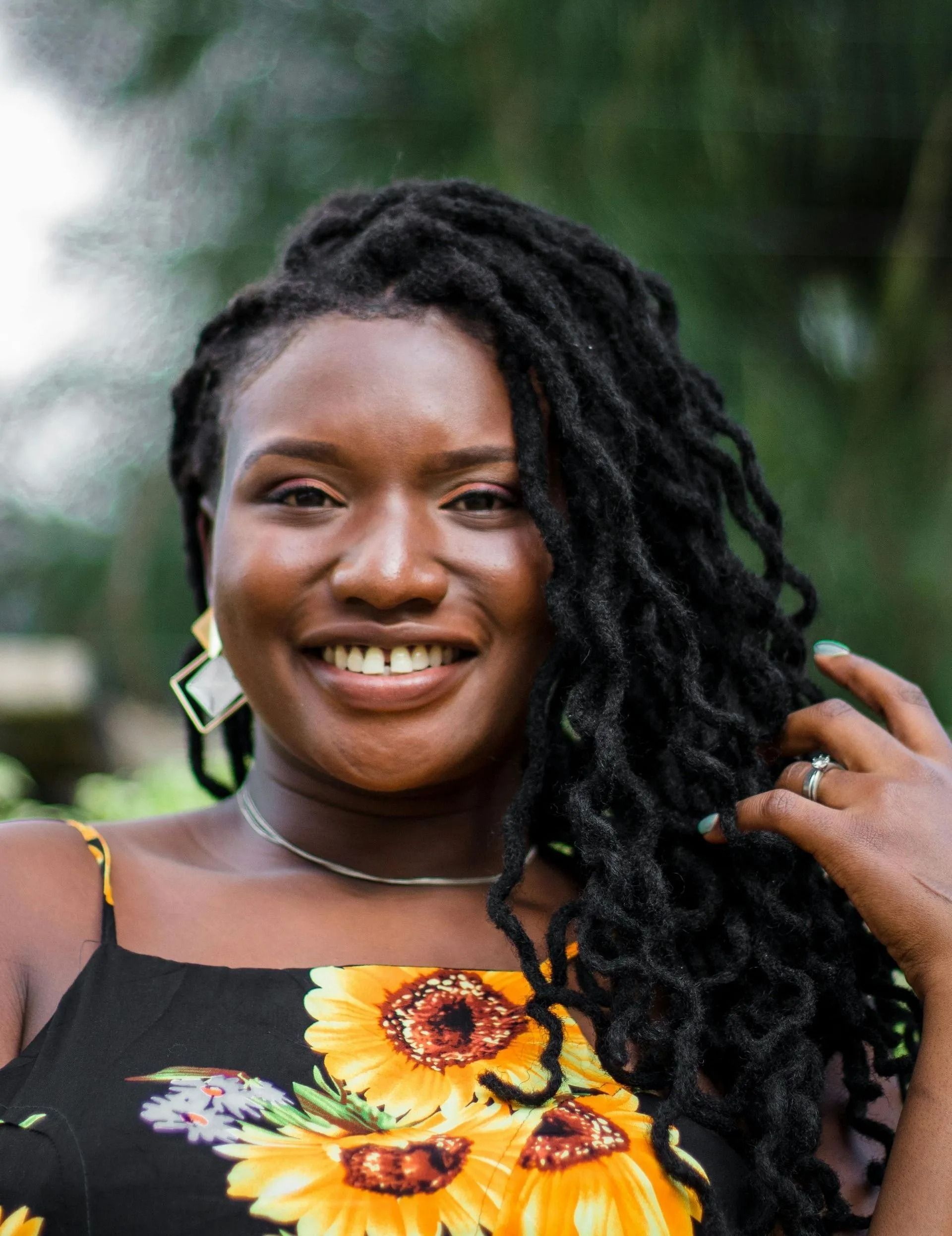 Smiling person with long dreadlocks in a black floral dress outdoors, touching their hair.