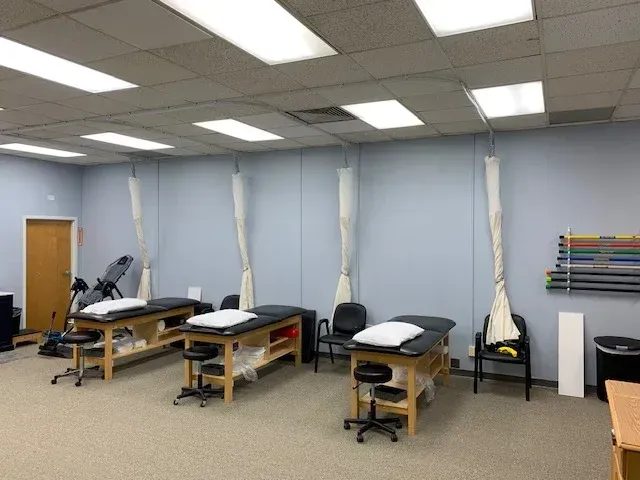 Physical therapy room with treatment tables, curtains, and exercise equipment. Light blue walls, beige carpet.