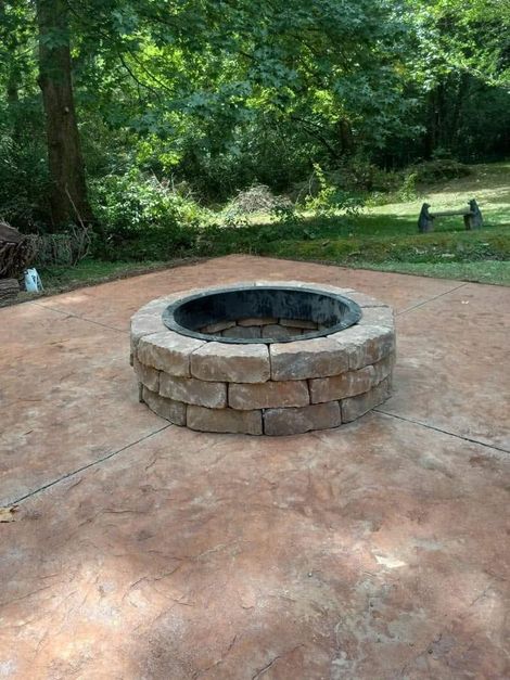Stone fire pit in a concrete patio surrounded by trees and greenery.