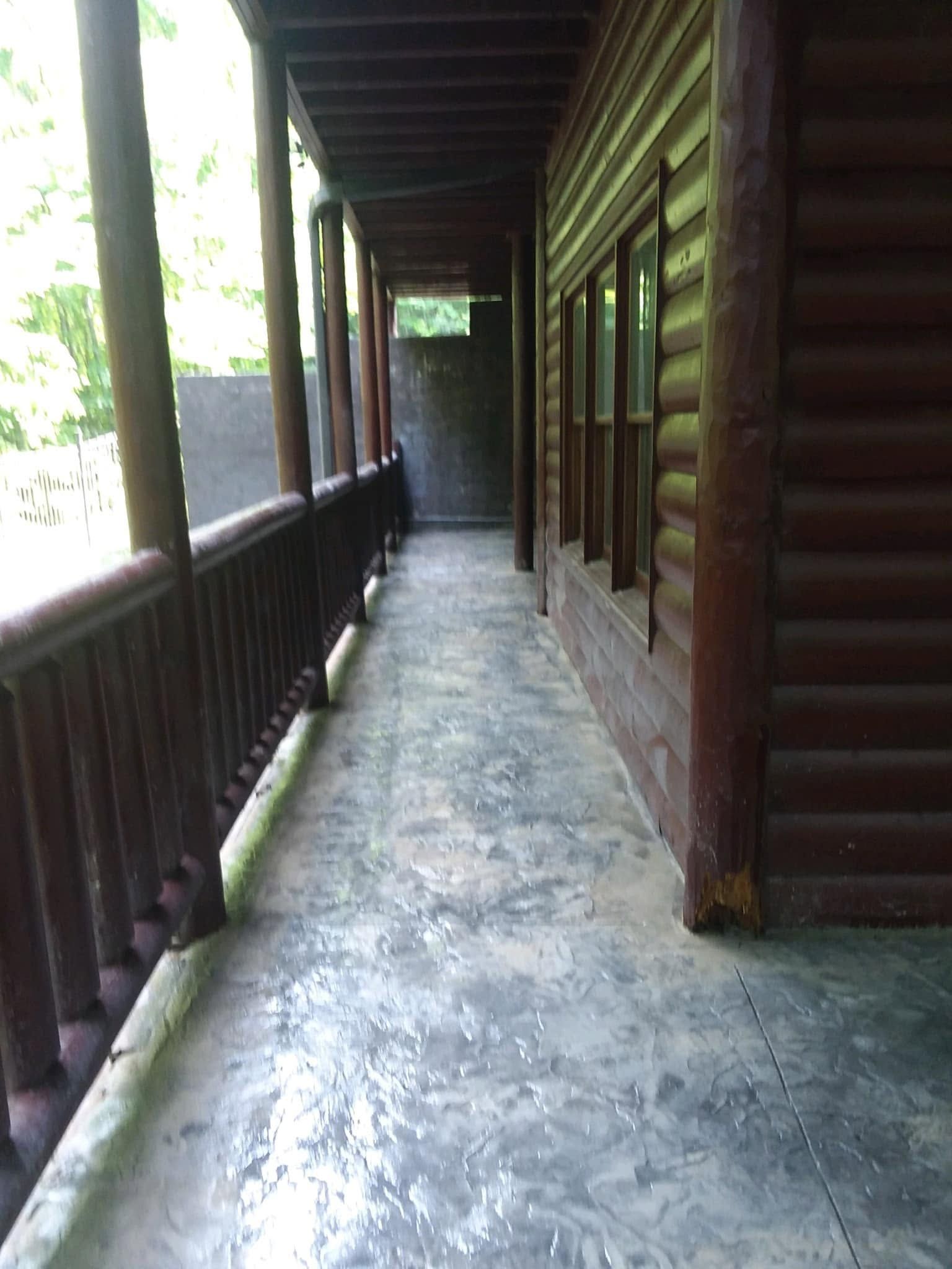 Wooden porch corridor with railings, log cabin walls, and a concrete floor, lit by daylight