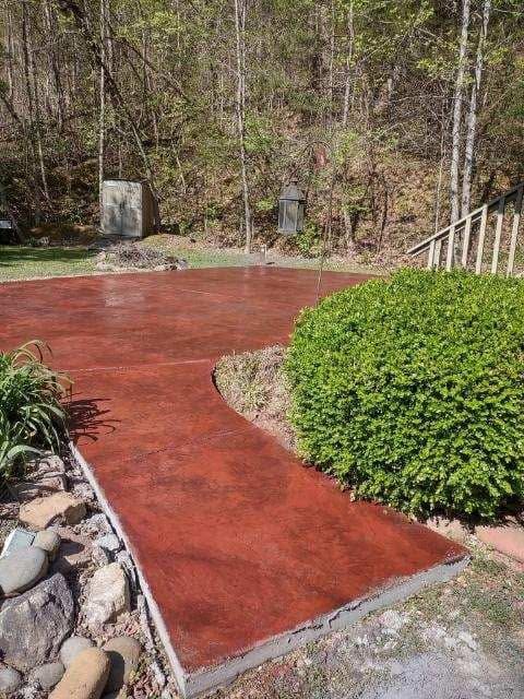 Red concrete patio with curved edge beside trimmed green shrub and stone border in a wooded backyard