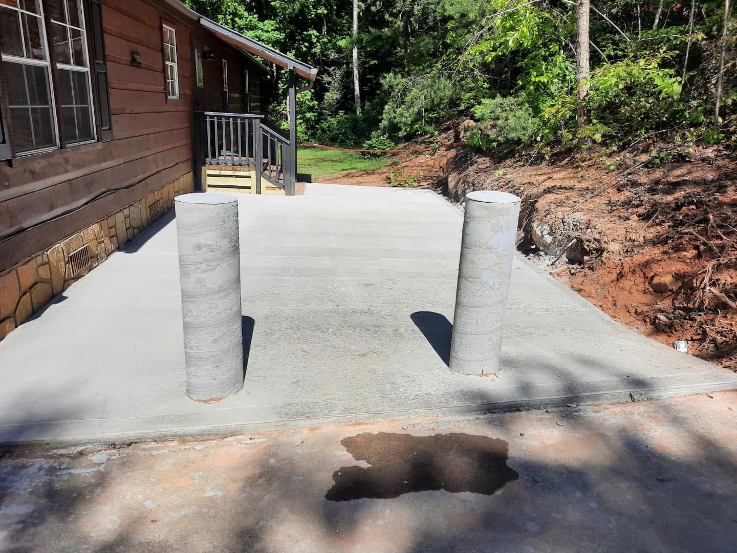 Concrete walkway with two short bollards beside a wooden building and a forested yard