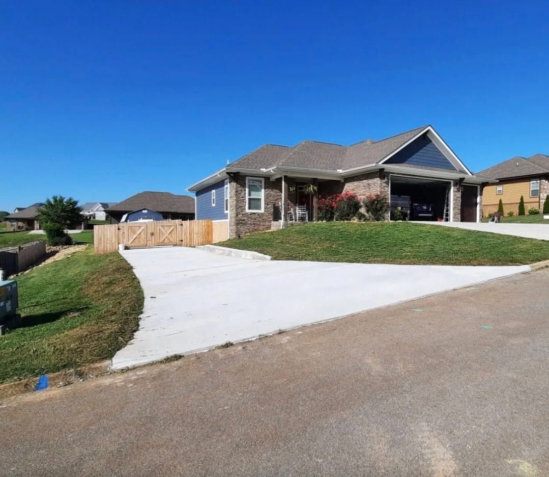 Single-story suburban house with a wide white driveway and green lawn under a clear blue sky