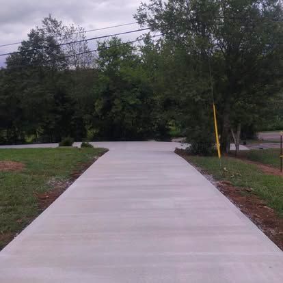 Concrete walkway leading into a wooded area beside a road under an overcast sky.