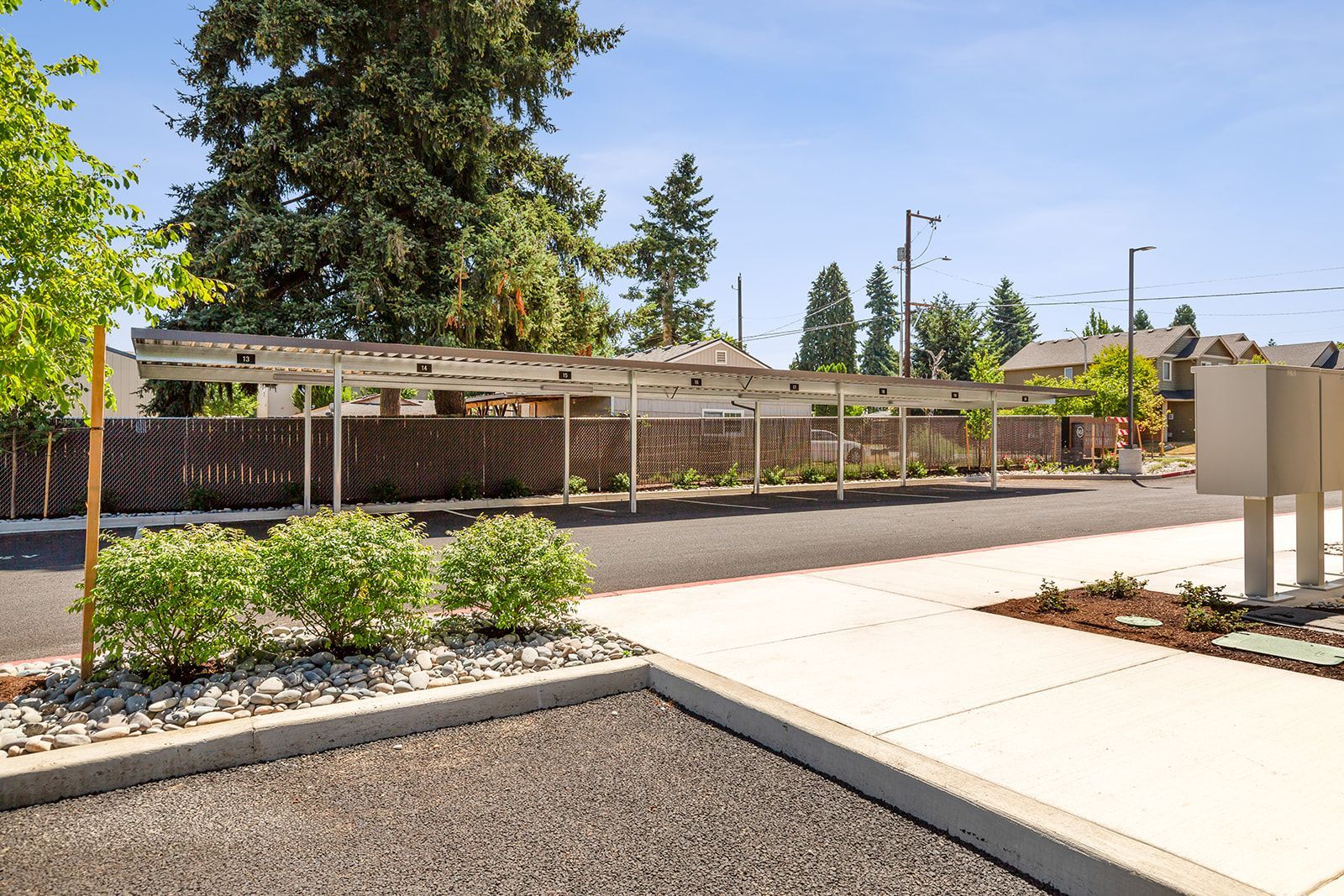 Outdoor walkway beside a fenced garden with shrubs, trees, and a bright blue sky
