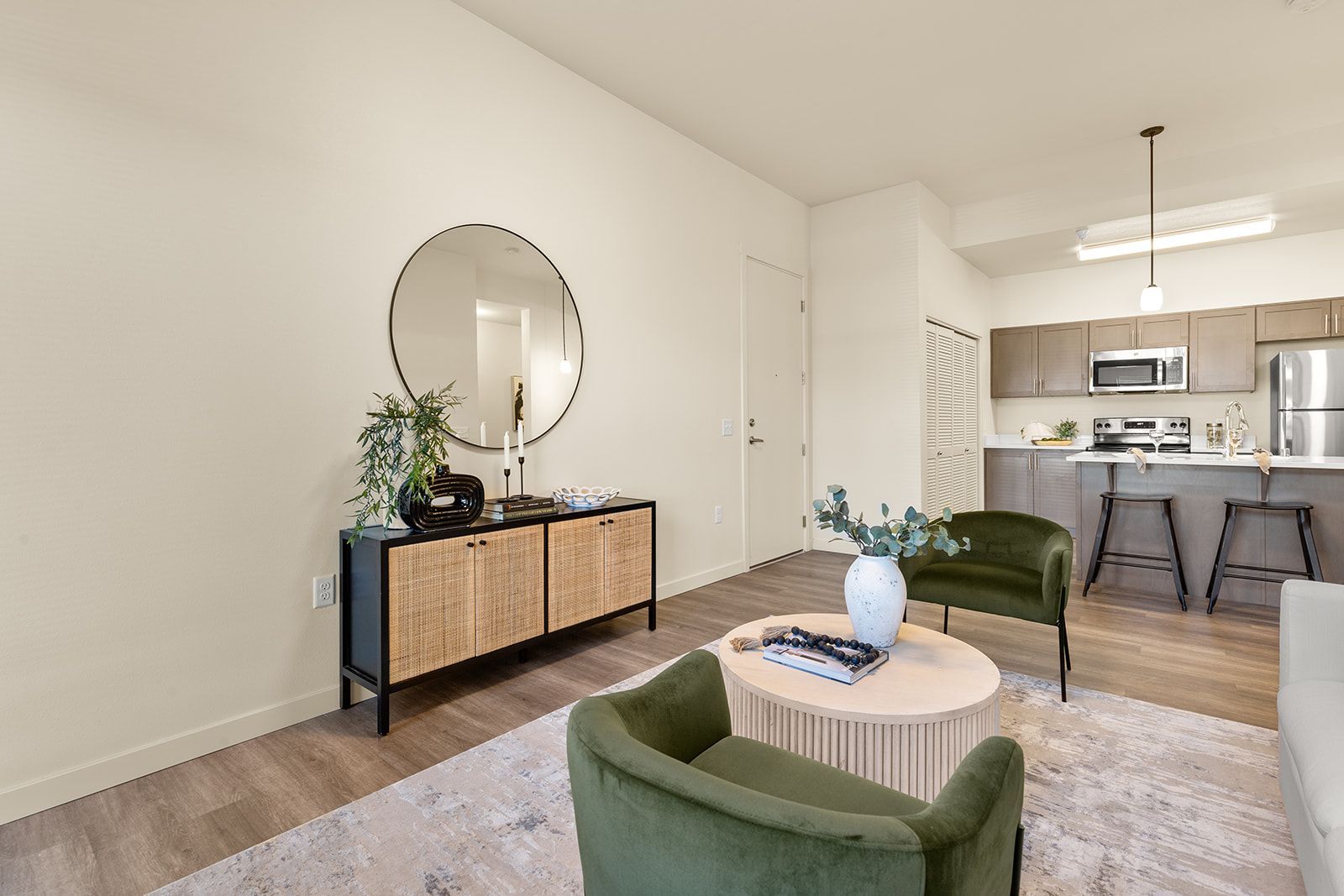 Bright living room with green chairs, round rug, sideboard, and an open view to a kitchen.