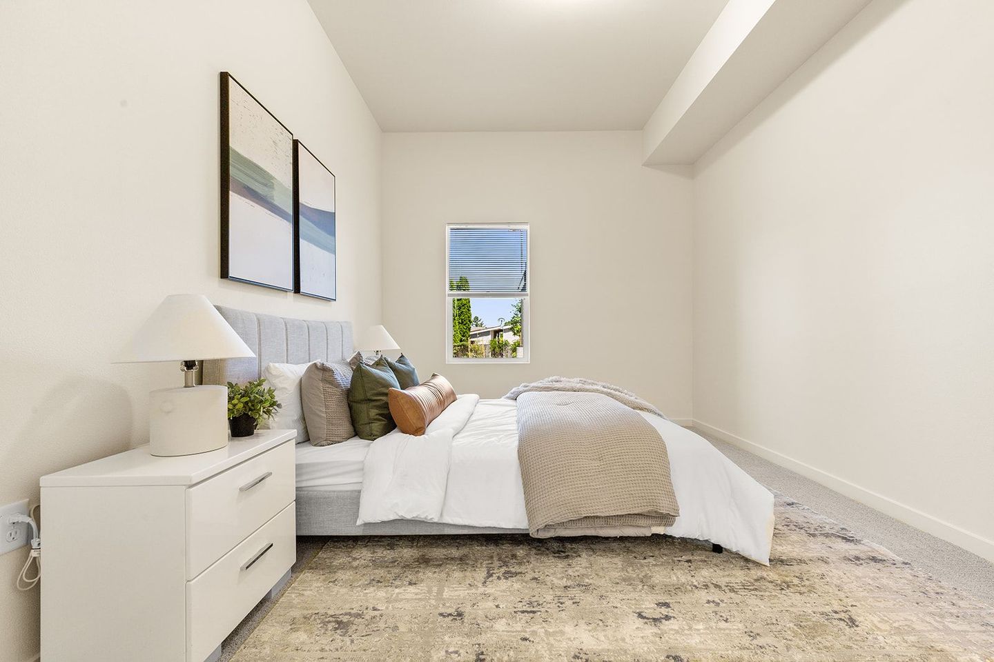 Bright minimalist bedroom with white bed, dresser, lamp, neutral decor, and small window.