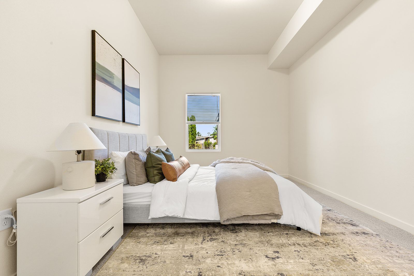 Bright bedroom with white bed, nightstand, lamp, artwork, and a small window.