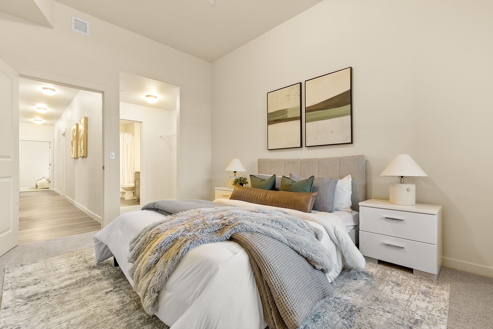 Bright bedroom with a bed, nightstands, lamps, wall art, and an open doorway to a hallway