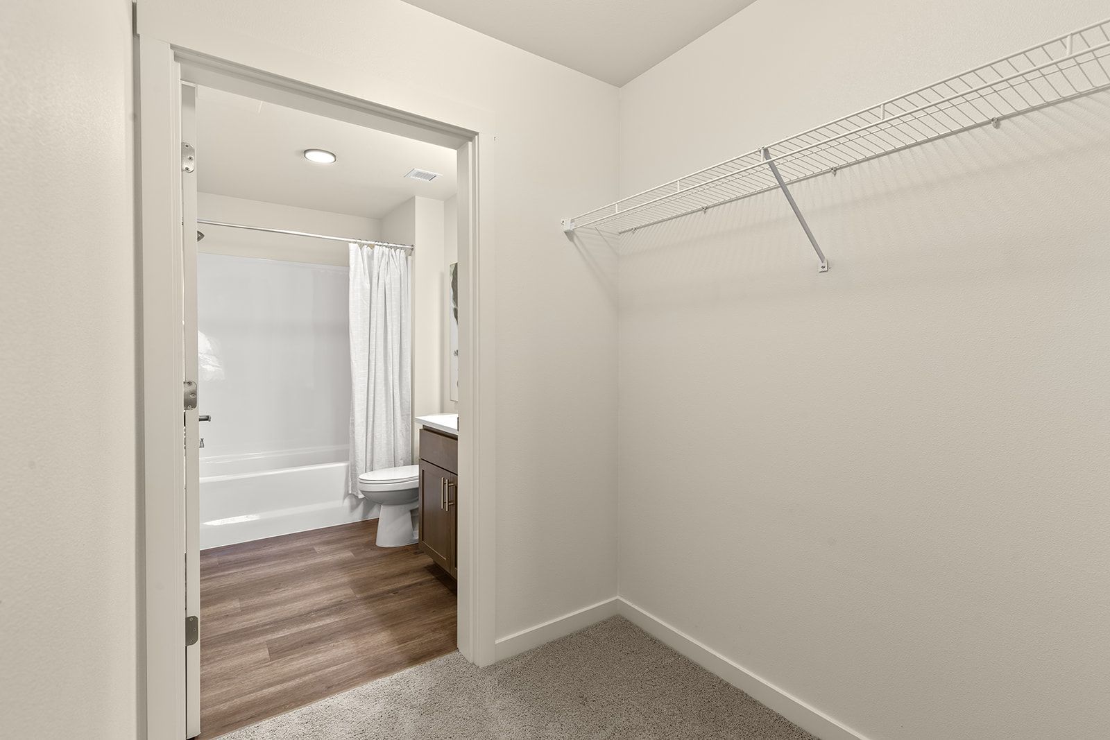 Empty carpeted walk-in closet with a wire shelf, open door, and view into a bathroom with a tub.