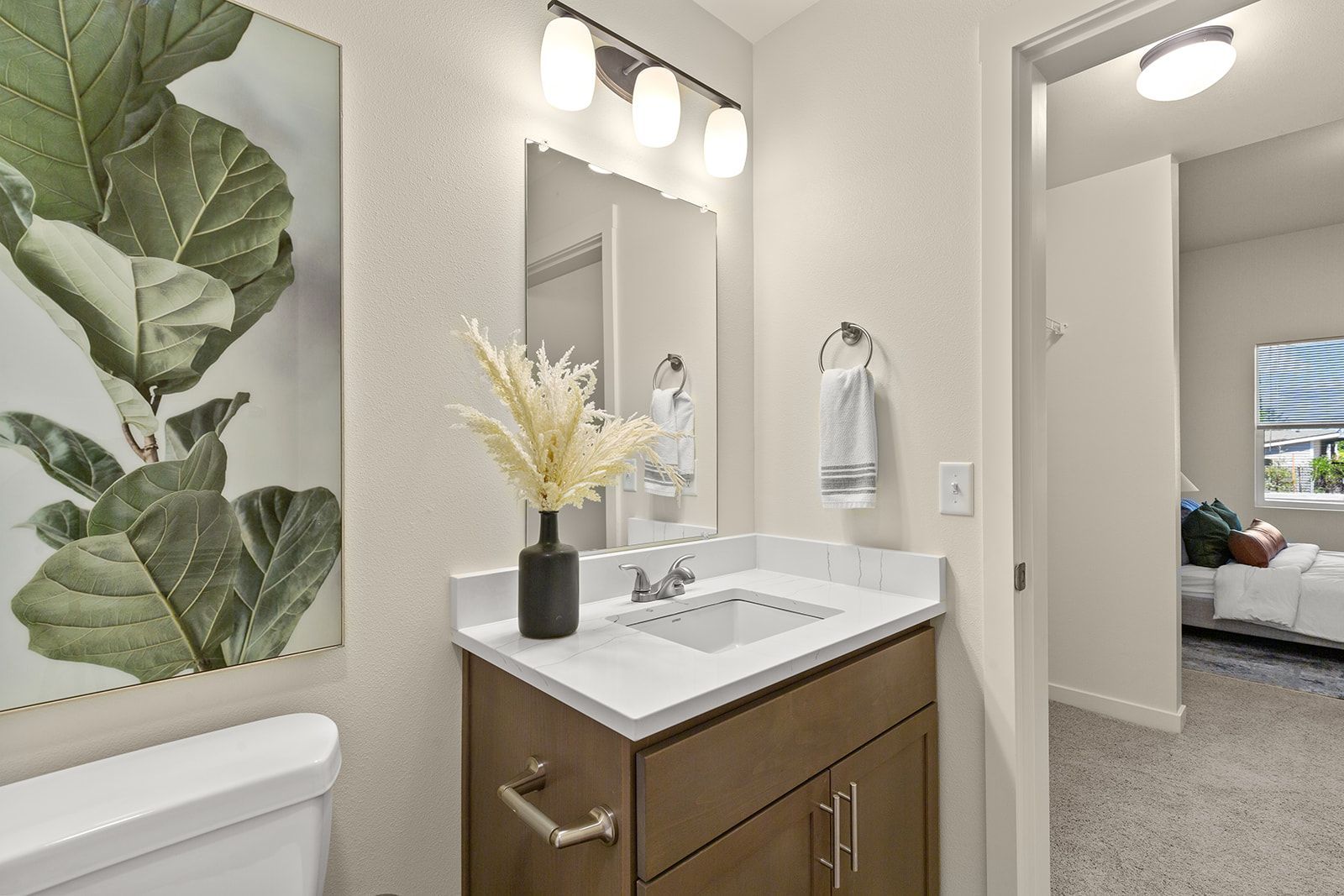 Bright bathroom vanity with mirror, sink, wall art, and a towel beside a bedroom doorway.