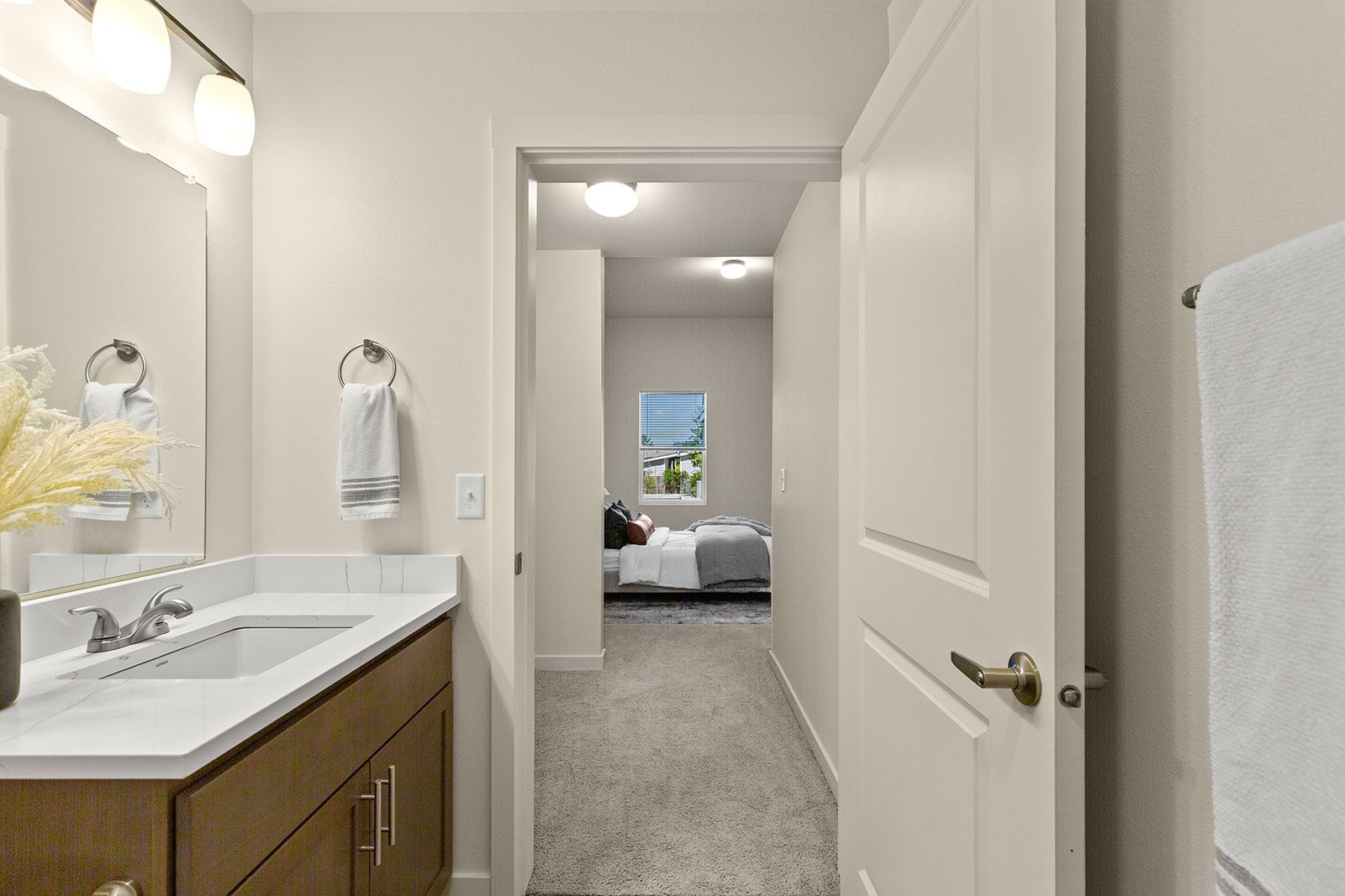 Bright hallway view from a bathroom toward a bedroom with white walls, carpet, and vanity sink.