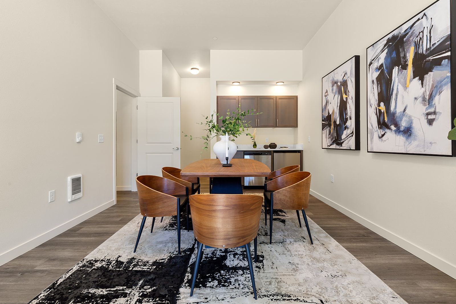Modern dining room with a wooden table, four chairs, abstract wall art, and a kitchen in the background