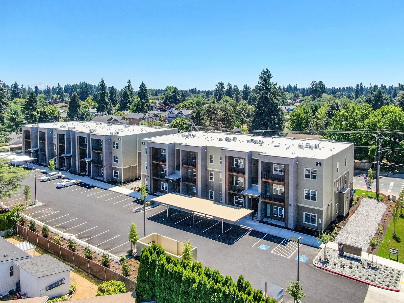 Aerial view of a modern apartment complex with parking lot, surrounded by trees under a clear blue sky