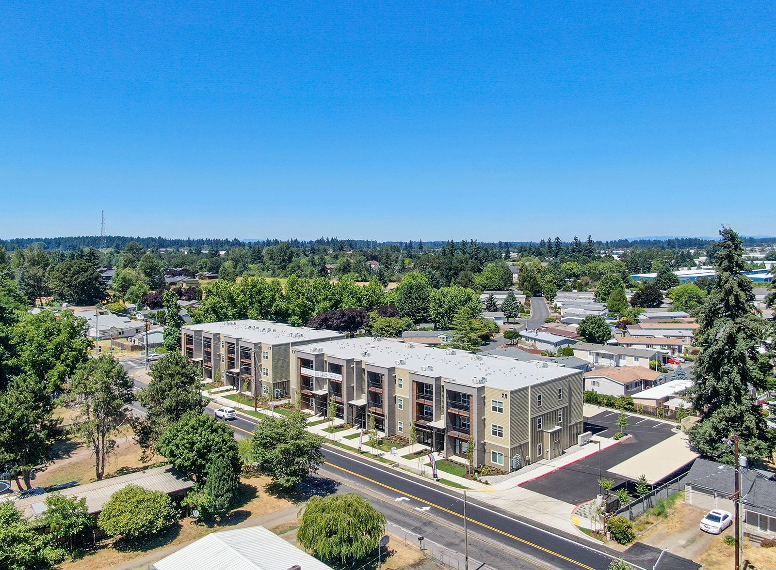 Aerial view of a modern apartment building along a road, surrounded by trees and low-rise homes under a clear blue sky