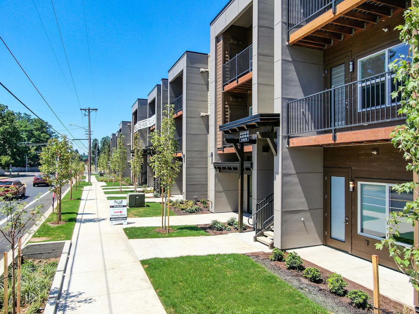 Modern apartment complex with balconies, sidewalk, trees, and landscaped lawn along a street.