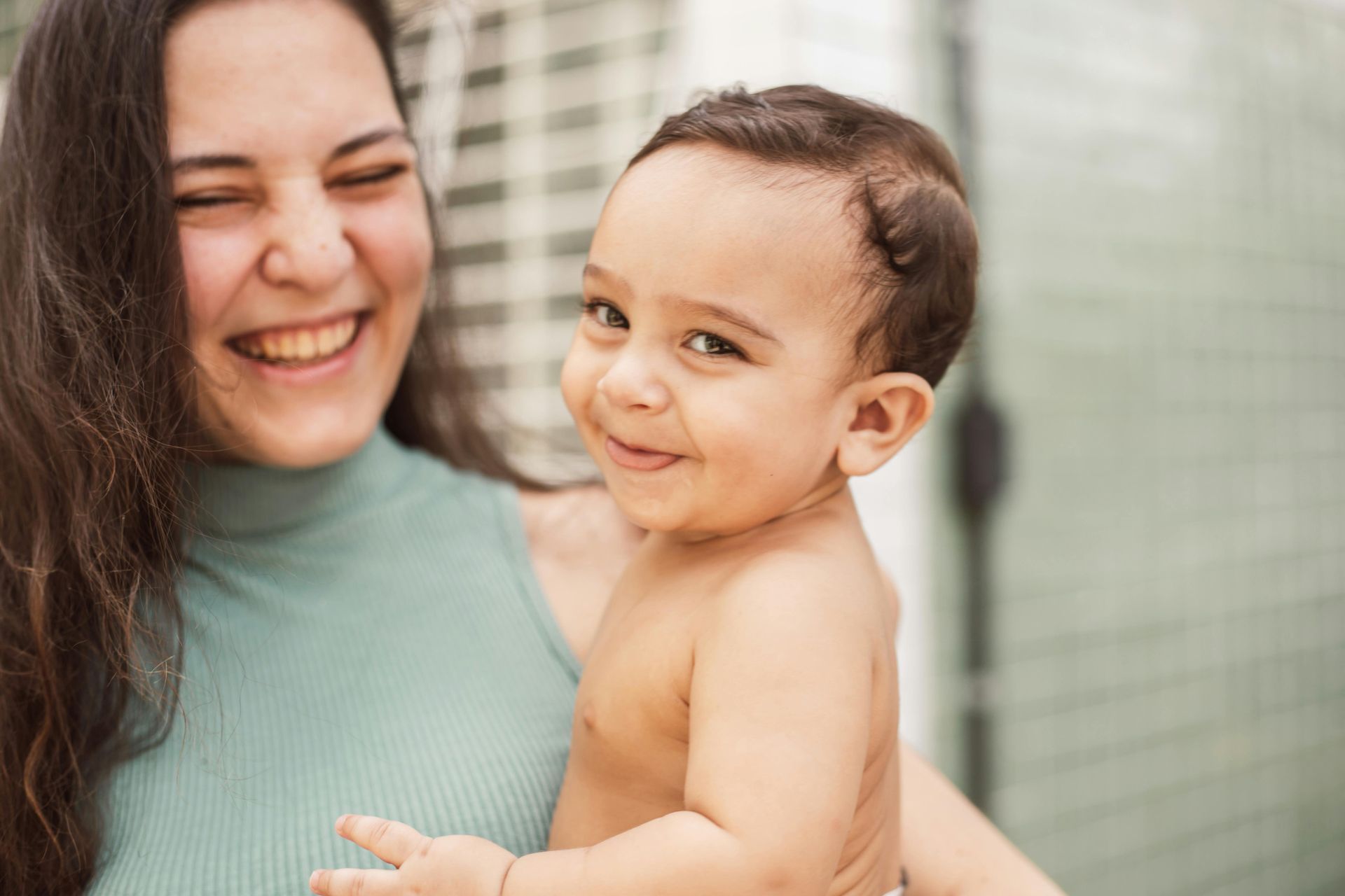 Woman smiling, holding a smiling baby. Both are indoors, baby is shirtless.