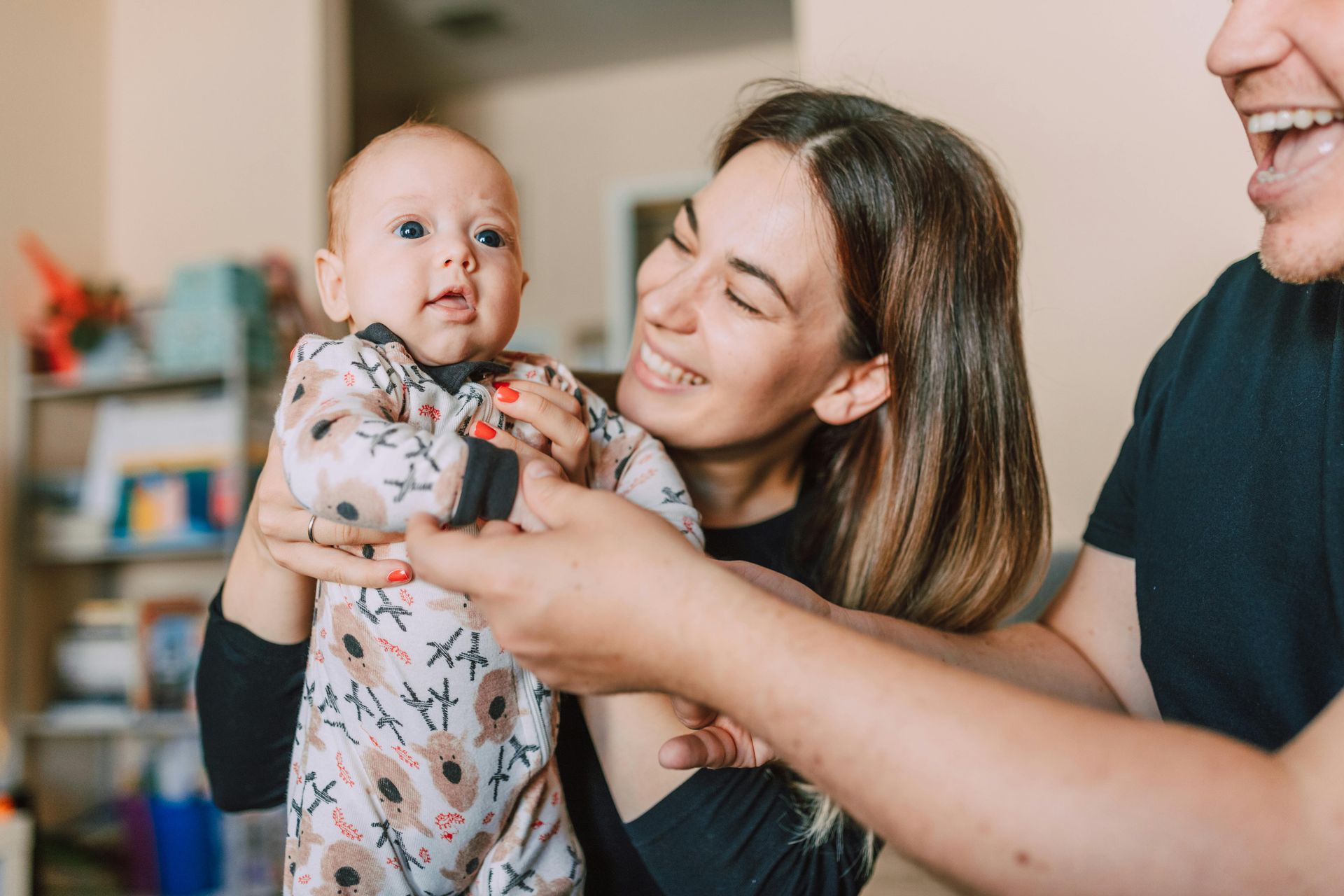 Woman holding baby, smiling, man beside them, laughing. Indoor setting.