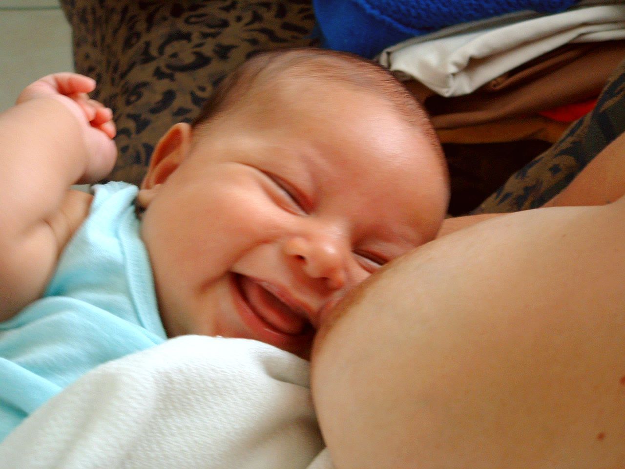 Smiling baby nursing, light blue onesie, next to a breast.
