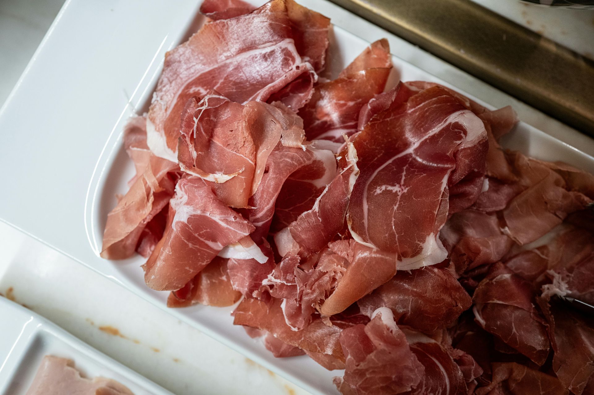 Thinly sliced prosciutto piled on a white tray, with pink-red marbled meat and visible fat edges