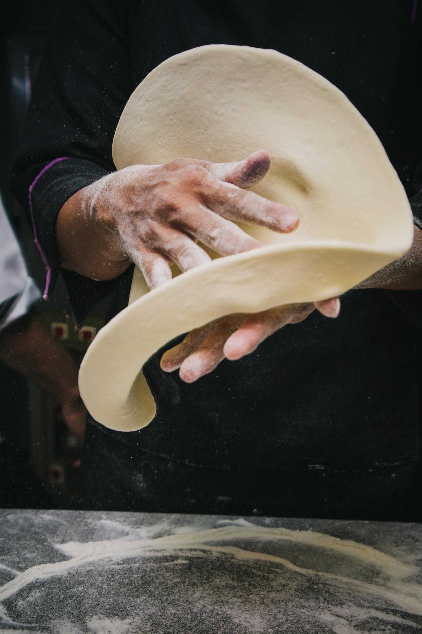 Hand stretching and tossing pizza dough against a dark background, with flour dust below