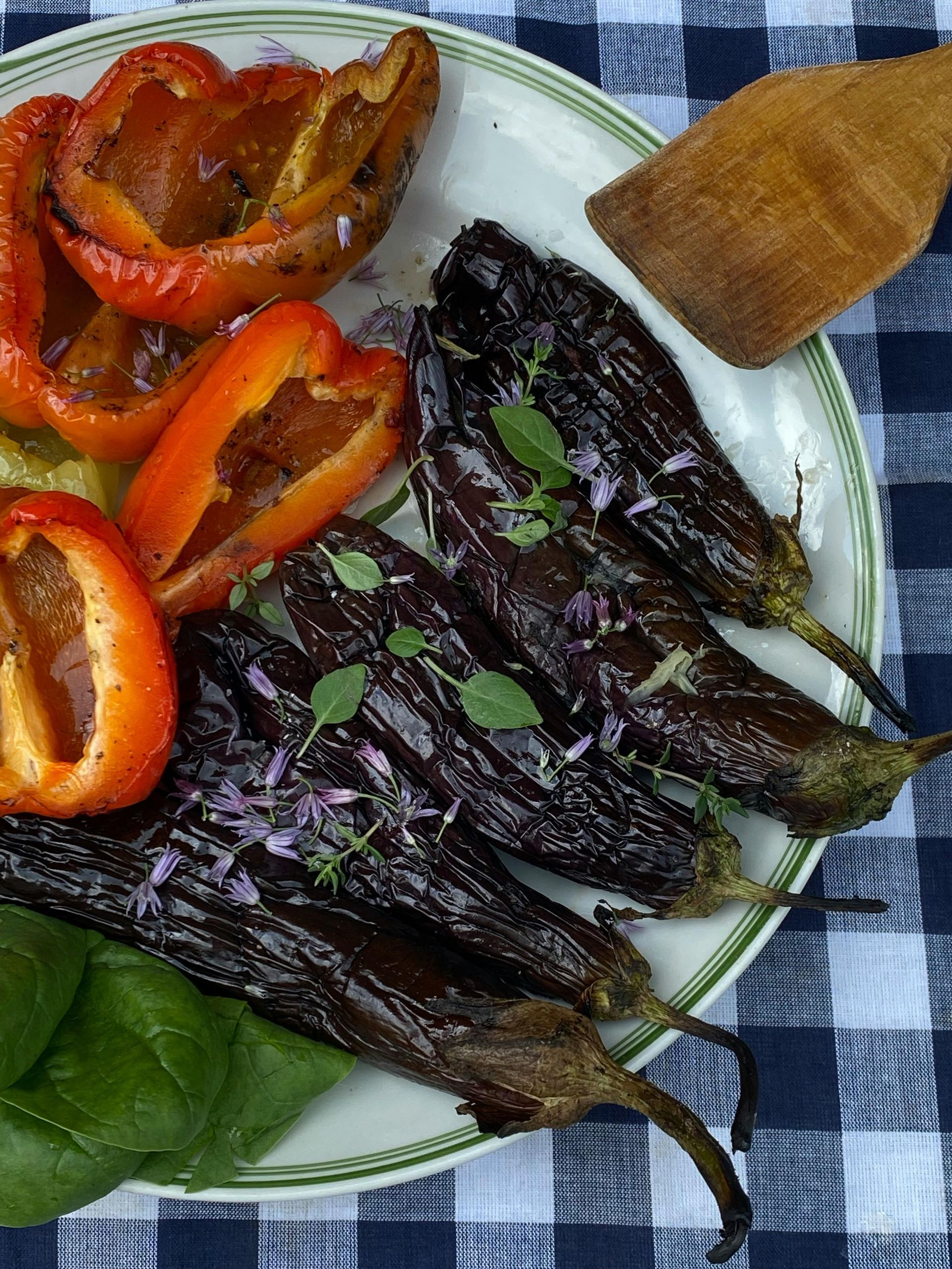 Roasted peppers and grilled dark vegetables on a plate with bread and greens on a checkered cloth.