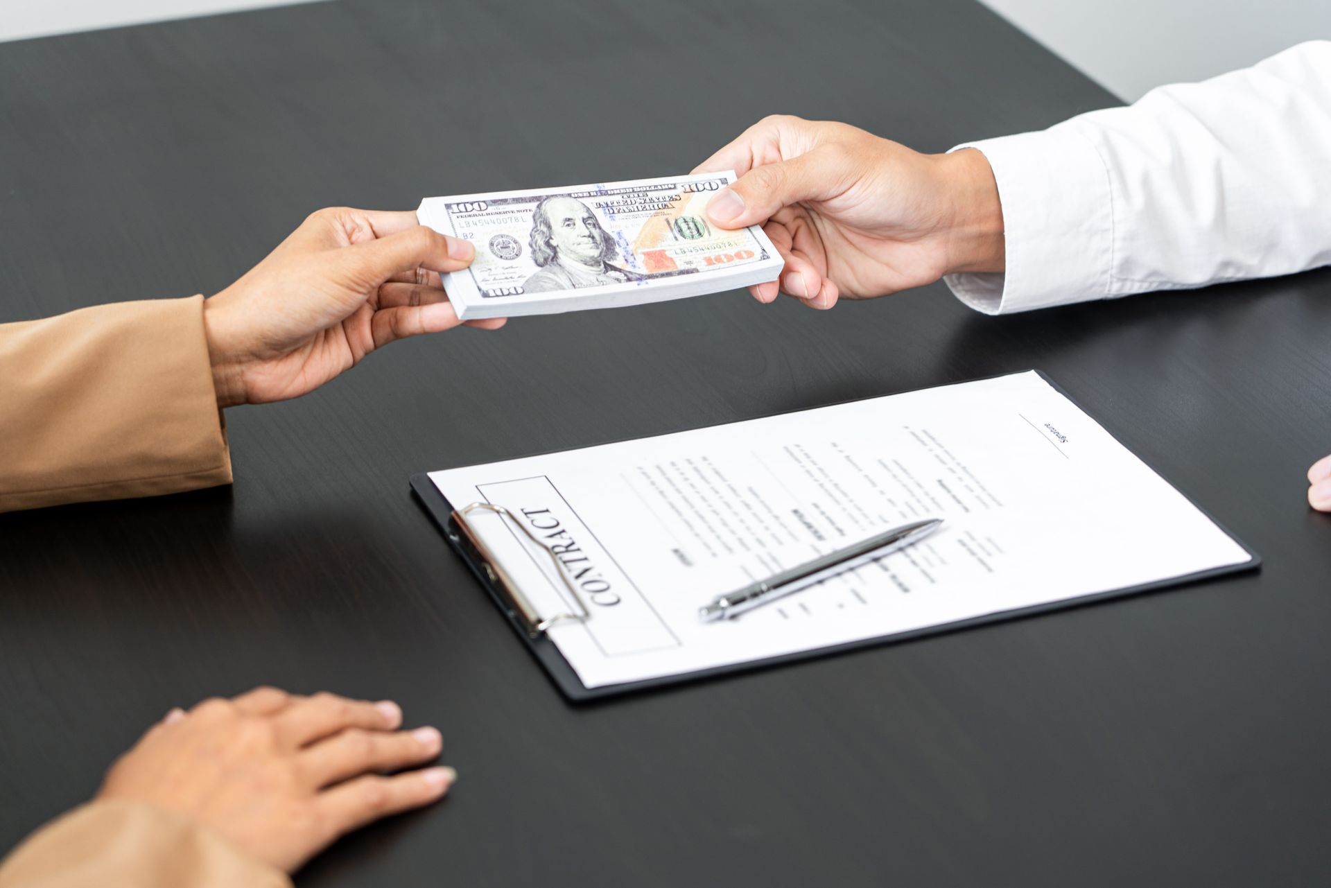Hands exchange cash over a desk with a signed document and pen.
