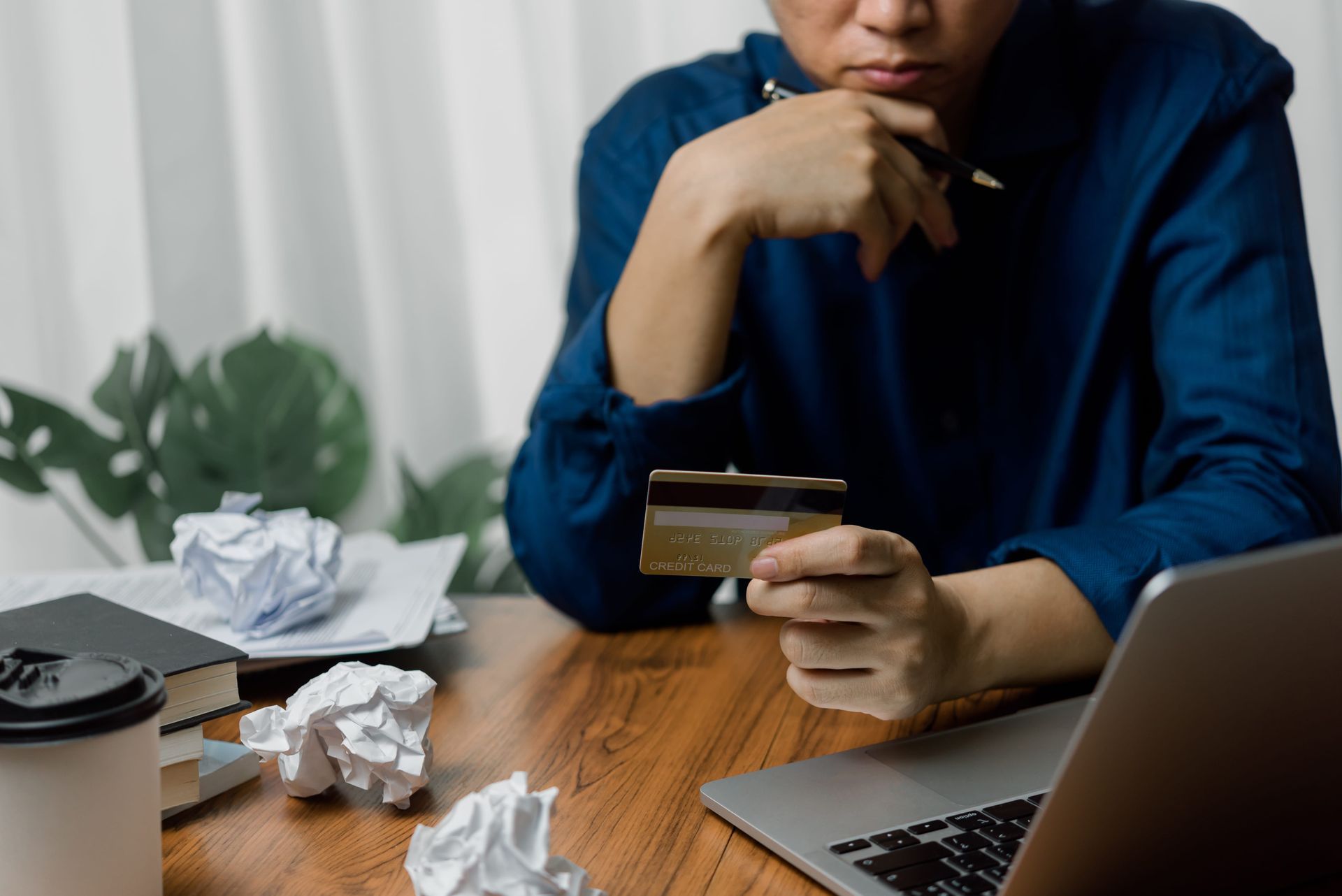 Person at a desk holding a credit card, with laptop, crumpled paper, and bills, looking stressed