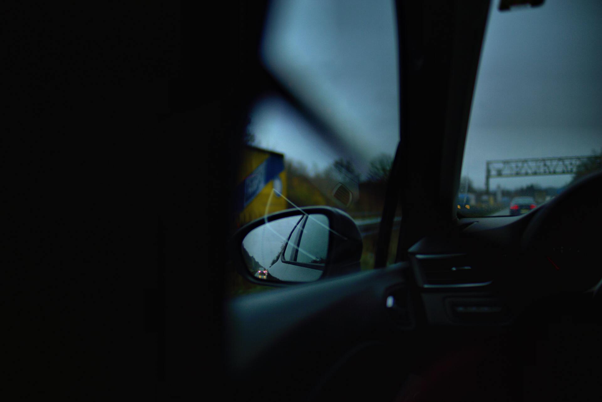 View from inside a car; side mirror reflecting a highway and overcast sky.