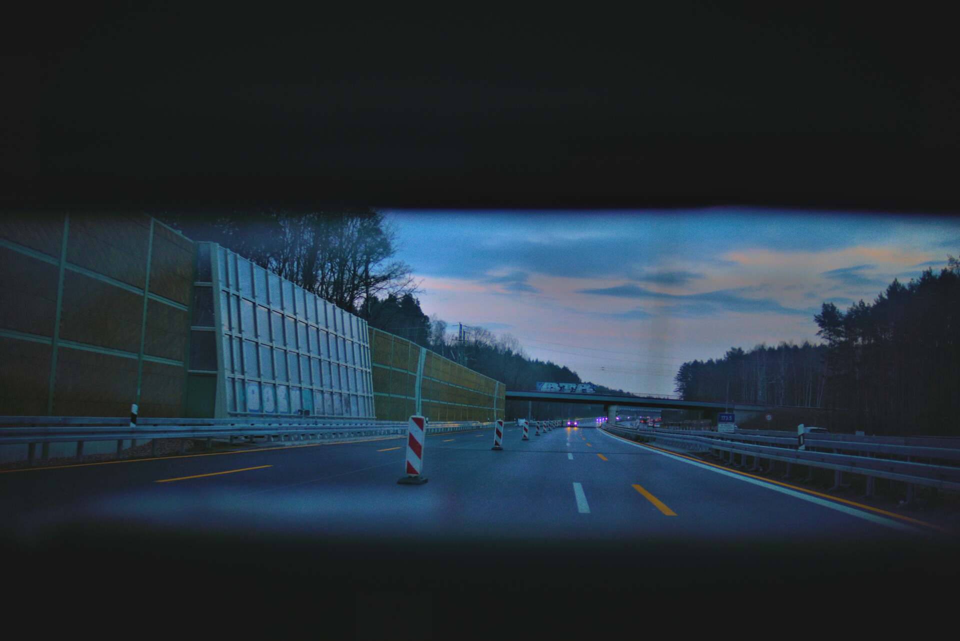 Highway view, roadway with sound barrier on the left, trees in the distance, dusk.