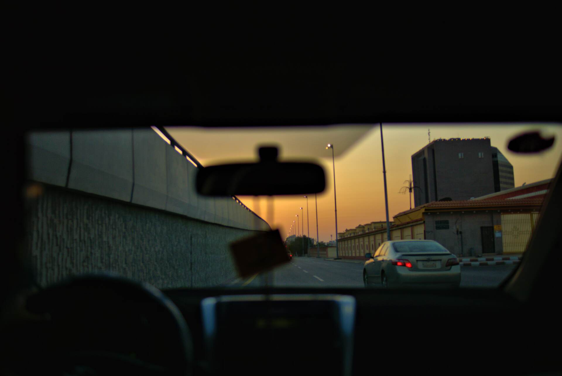 View from inside a car at sunset, road ahead with other vehicles, buildings in the distance, and a sunlit sky.