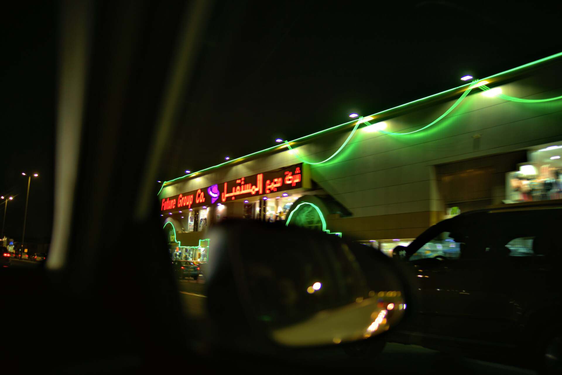 Exterior of a store at night, illuminated by green lights. Arabic text is visible above the entrance.