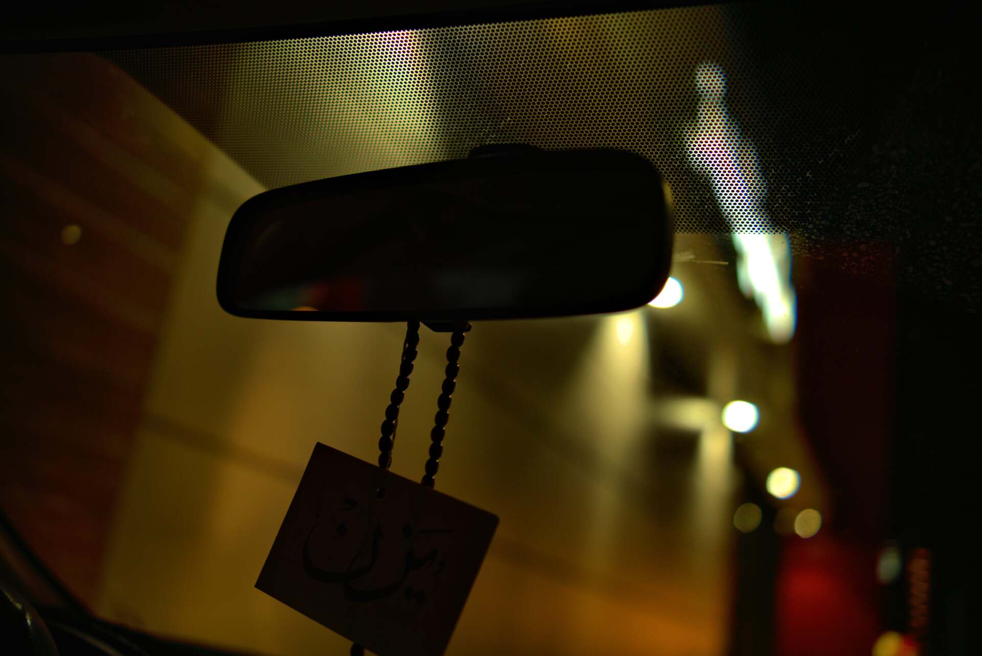 Interior car view, rear-view mirror with dangling air freshener; lights blur in background.