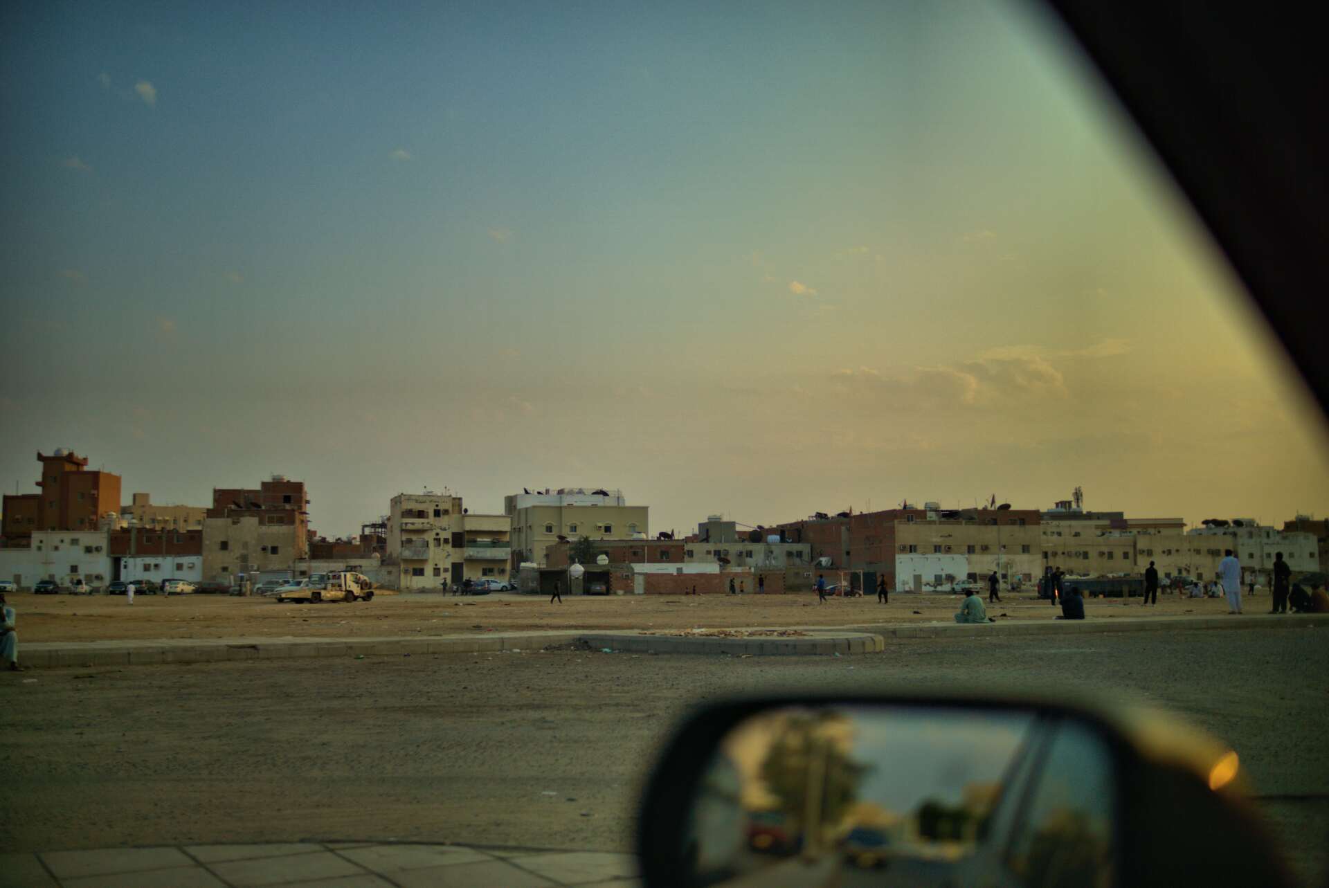 Buildings and people in a desert setting at dusk, seen from a car window, with a side mirror in view.