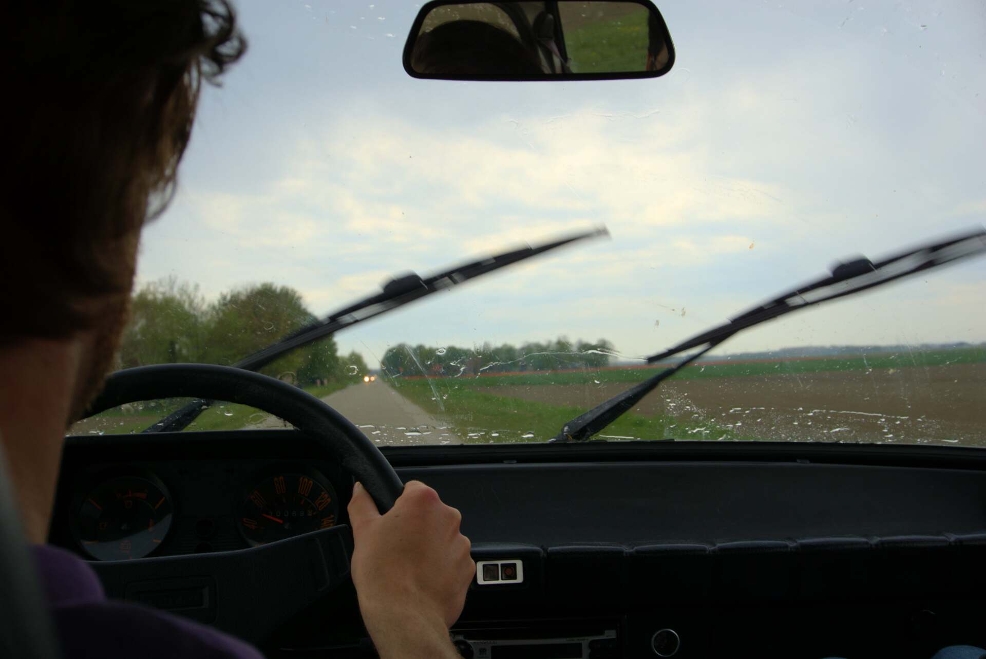 Person driving a car, windshield wipers on, view of road and field.