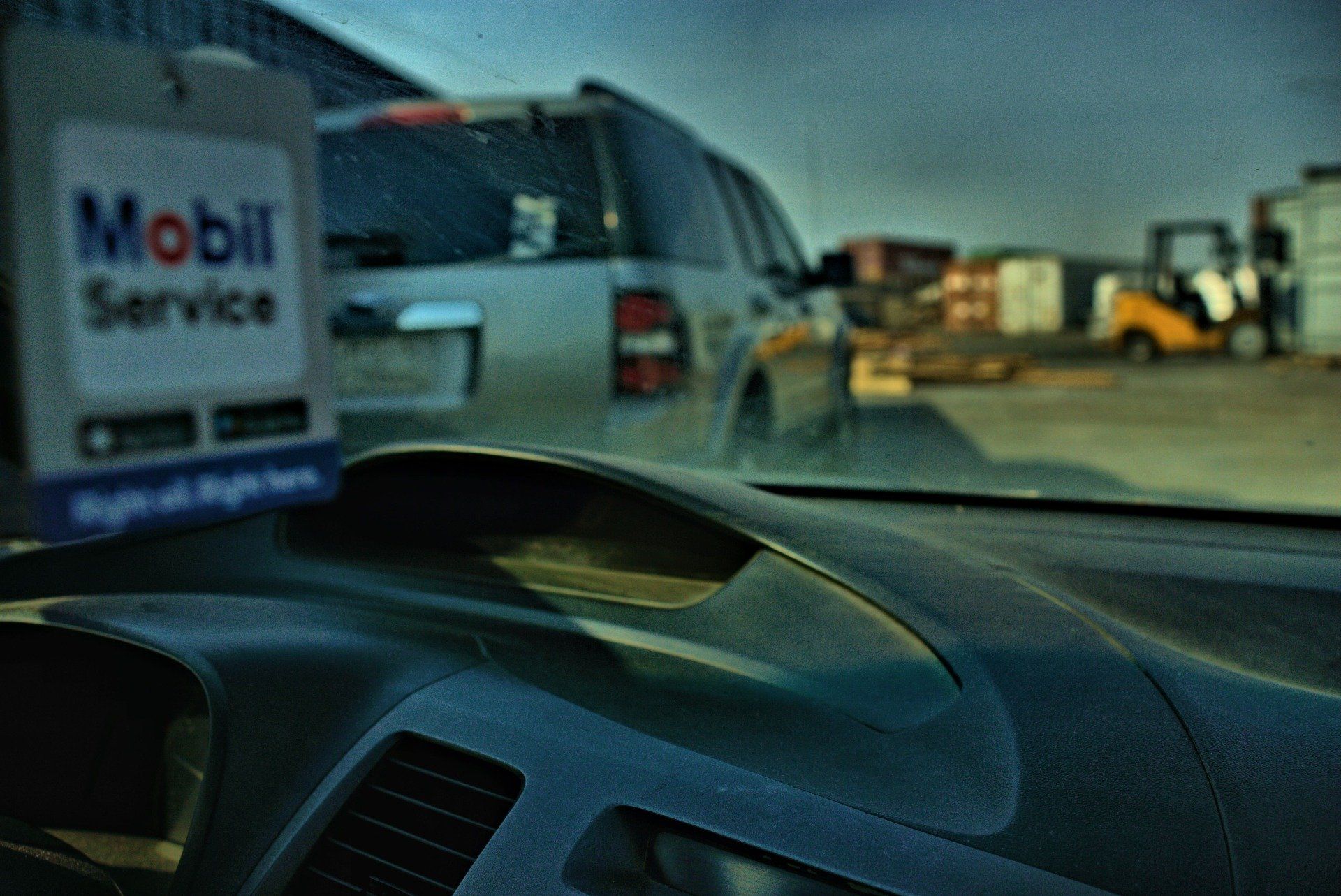 Dashboard view with Mobil Service air freshener, looking out at a silver SUV and industrial setting.