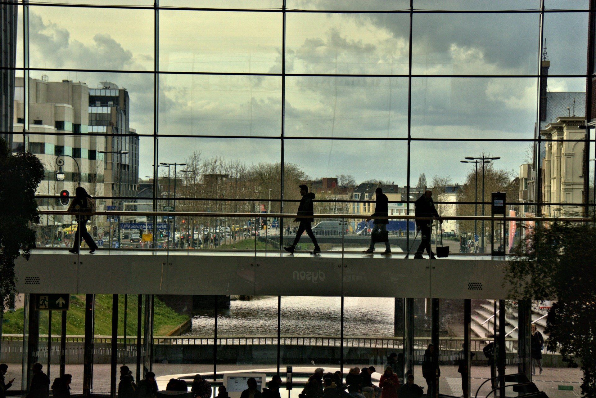 Silhouetted figures walk across a walkway, viewed through a large window. Buildings and a river are visible outside.