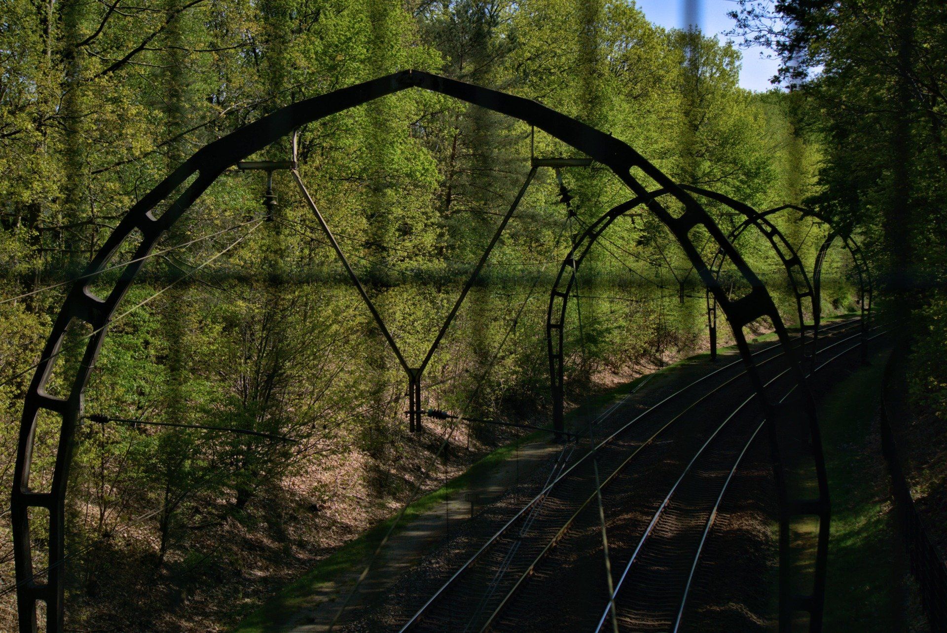 Train tracks passing under arched metal structures, surrounded by trees.