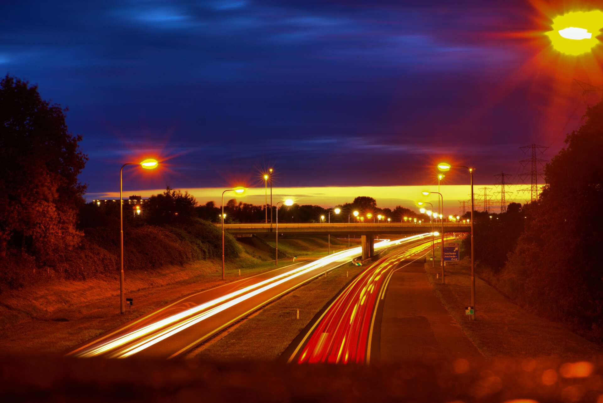 Highway at dusk with streaks of car lights under streetlights and a dark blue sky.