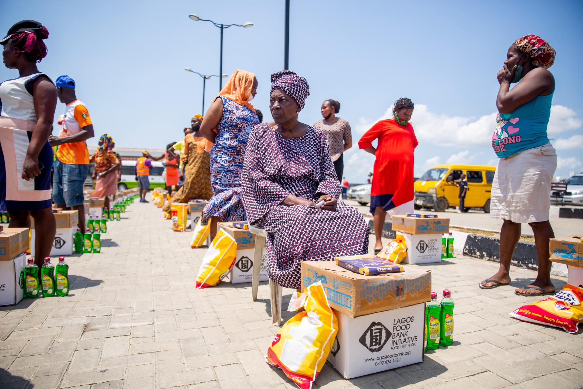 People waiting for aid, sitting and standing near boxes and bags on a paved area; sunny day.