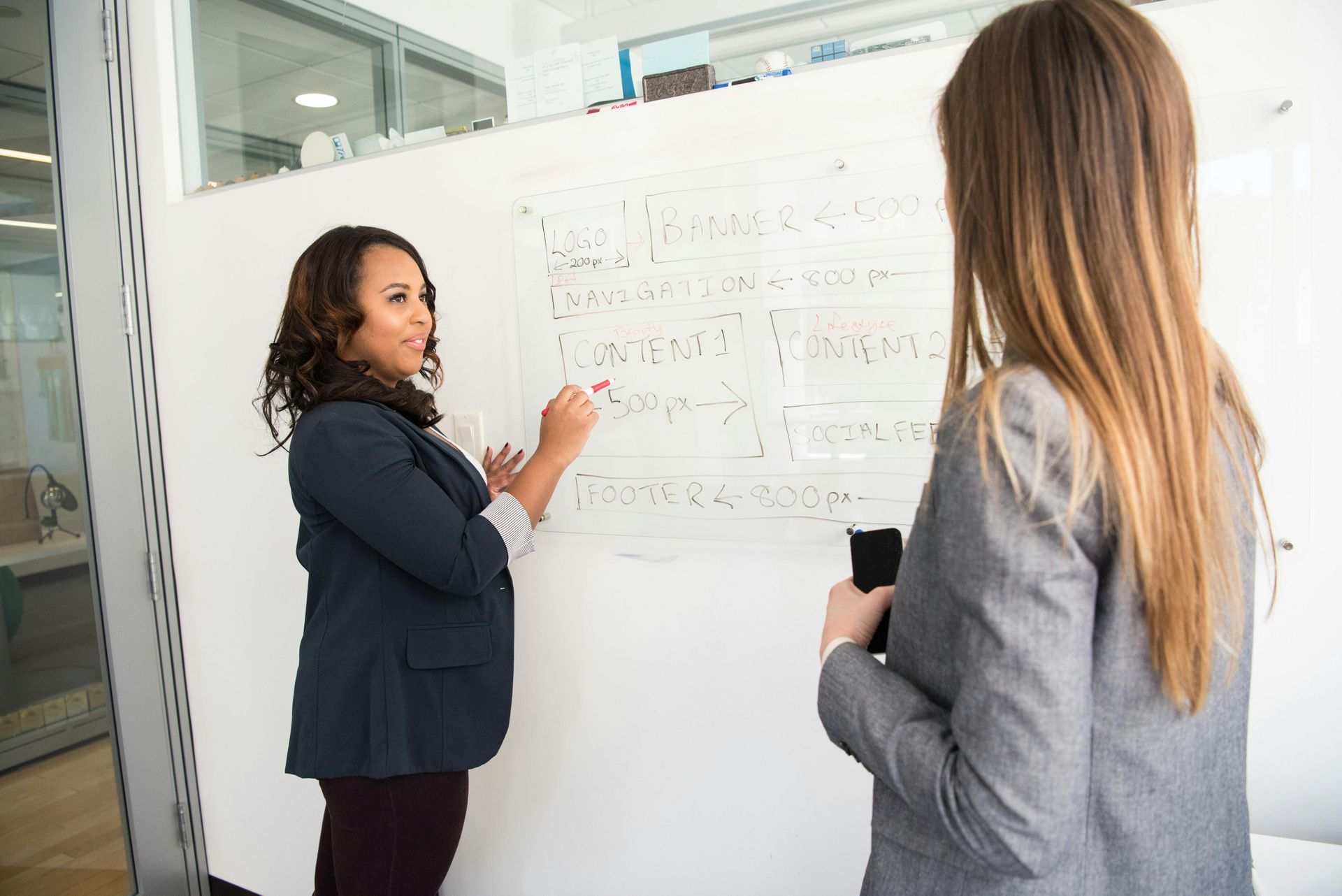 Woman in blazer writes on whiteboard as another woman watches, both in an office setting.