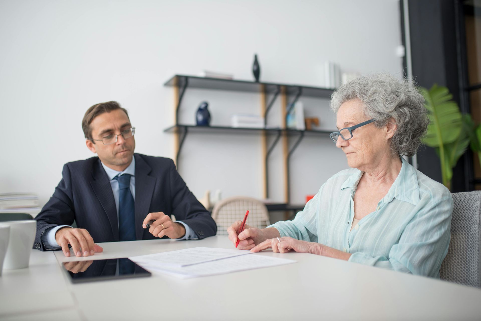 Man in suit looks on as woman signs a document at a desk in an office setting.